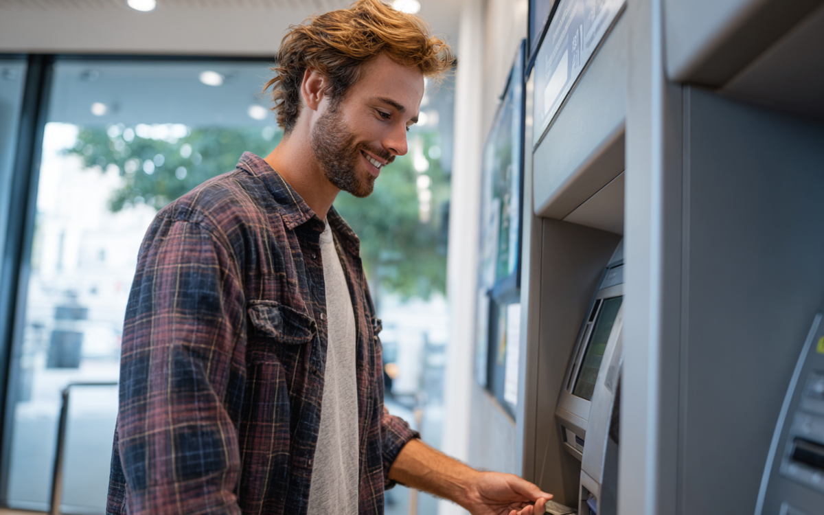 Junger Mann bedient im Freien einen Geldautomaten und zieht mit seiner Karte Bargeld.